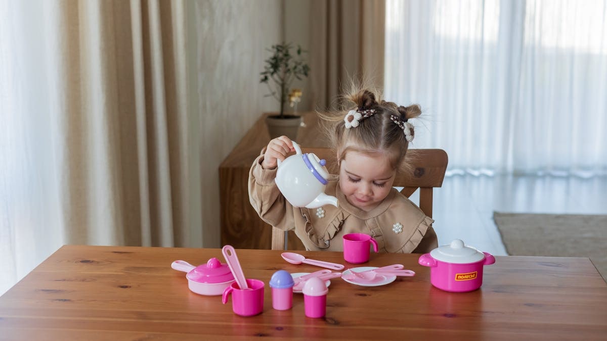Cute young girl having fun with a toy tea set on a wooden table indoors.